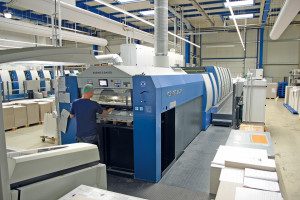 Zu sehen ist eine unserer Offsetdruckmaschine in die Flucht fotografiert. An der Auslage steht ein Medientechnologe Drück mit dem Rücken zugewandt.
